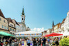 Großer Stadtplatz mit Häusern, Kirche, viele Menschen um Verkaufsstände und sitzend auf Bierbänken