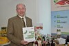 Landesrat Max Hiegelsberger mit einem Buch in der Hand, im Hintergrund ein Tisch mit verschiedenen regionalen Produkten, Flaschen und Gläser. Auf dem Buch steht der Titel Wie’s Innviertel schmeckt