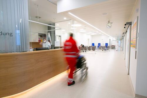 Ein Sanitäter ist von hinten zu sehen, der jemanden im Rollstuhl an der Anmeldung im Eingangsbereich eines Krankenhauses vorbeischiebt. 