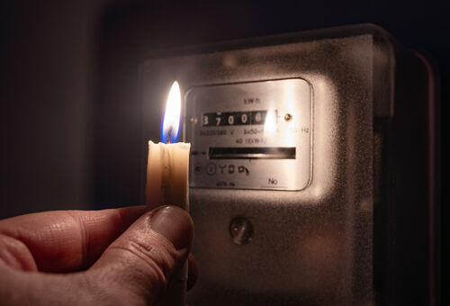 Hand hält eine brennende Kerze im Dunkeln zu einem Stromzähler, um die Daten ablesen zu können.