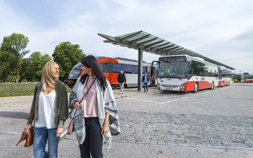 Zwei junge Frauen gehen nebeneinander und unterhalten sich; im Hintergrund sind ein Zug und mehrere Busse zu sehen. 