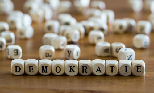 Auf einem Tisch liegen Holzwürfel mit Buchstaben, im Vordergrund wird daraus das Wort Demokratie gebildet