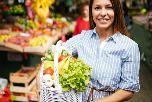 Frau vor einem Marktstand hält einen Einkaufskorb gefüllt mit Gemüse und Obst