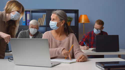 Lehrerin mit Maske hilft einer erwachsenen Frau am Laptop, im Hintergrund ein jüngerer und ein älterer Mann, der Jüngere schreibt etwas auf einen Notizblock.