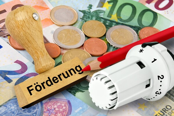 Euro-Scheine und Euro-Münzen, darauf liegen ein Stempel mit der Aufschrift ‚Förderung‘, ein Rotstift und ein Heizkörper-Thermostatkopf. 
