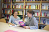 zwei Frauen sitzen nebeneinander an einem Tisch und unterhalten sich; eine von ihnen hält eine Broschüre in Händen, hinter ihnen ist ein großes Bücherregal zu sehen