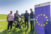 Jiří Falout, Landesrat Markus Achleitner auf einem Fahrrad, Edwin Sternad und Ing. Markus Obermüller stehen nebeneinander auf einer Wiese, neben ihnen ein Rollplakat mit dem Europa-Sternkreis und Beschriftung Interreg, Österreich und Tschechische Republik