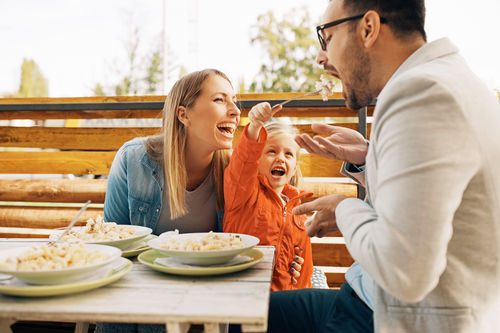  Mutter, Vater und die kleine Tochter essen gut gelaunt Pasta auf dem Balkon.