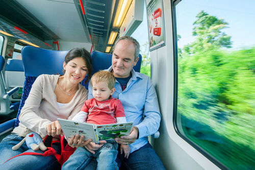 Familie sitzt im fahrenden Zug, die Eltern lesen einem kleinen Kind aus einem Buch vor.