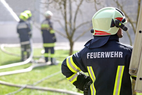 Ein Feuerwehrmann steht neben einer Leiter, im Hintergrund drei weitere Feuerwehrleute, die einen Feuerwehrschlauch halten, aus dem Wasser spritzt