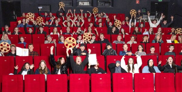 Jugendliche sitzen in einem Kinosaal, einige halten Preise in die Höhe. Aufnahme vom Filmpreis DEX 2024. 