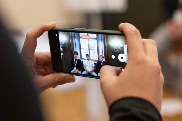 Hände halten ein Smartphone und filmen drei Jugendliche bei einer Diskussion.
