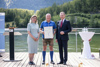 Cristina Biasetto, Ing. Johann Immervoll mit Urkunde und Landeshauptmann Mag. Thomas Stelzer auf einem Steg am See, im Hintergrund Wasser, Berge, Wald