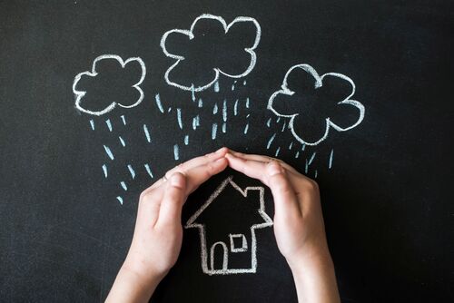 Hände halten sich schützend über ein auf eine Tafel mit Kreide gezeichnetes Haus, über dem Regenwolken stehen