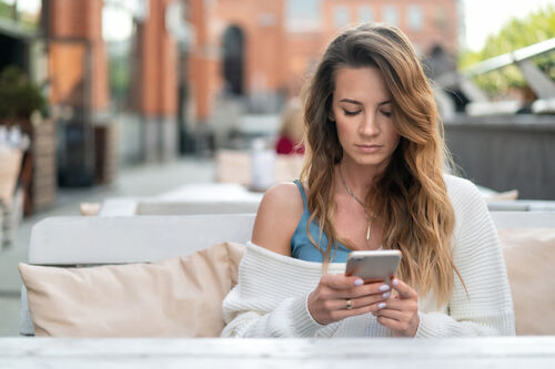 Eine junge Frau sitzt allein in einem Gastgarten und schickt mit ihrem Handy eine Mail an die Frauenberatung. 