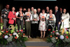 Gruppenbild mit vierzehn Personen stehend in zwei Reihen auf einer blumengeschmückten Bühne