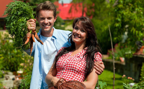 Eine Frau und ein Mann in einem Garten, der Mann hält einen Bund Karotten hoch
