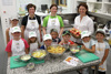 Acht Kinder und drei Erwachsene, alle mit Kochschürzen, unter ihnen Landesrätin Michaela Langer-Weninger, stehen in einer Großküche um einen Arbeitstisch, auf dem sich verschiedene Lebensmittel in Schüsseln befinden