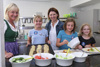 Drei Kinder sowie eine weitere Person und Landesrätin Michaela Langer-Weninger stehen in einer Großküche um einen Arbeitstisch, auf dem sich verschiedenen Lebensmittel in Schüsseln, auf einem Backblech und auf Tellern befinden