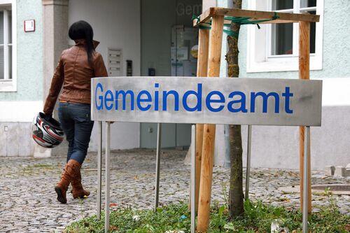 Frau geht zu einem Hauseingang, davor steht ein Schild mit der Aufschrift „Gemeindeamt“