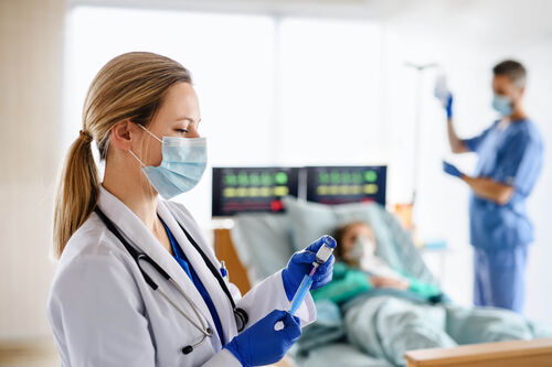 Ärztin in einem Krankenzimmer zieht eine Spritze auf, im Hintergrund Krankenhausbett mit Patient und Pflegekraft, die Infusion vorbereitet