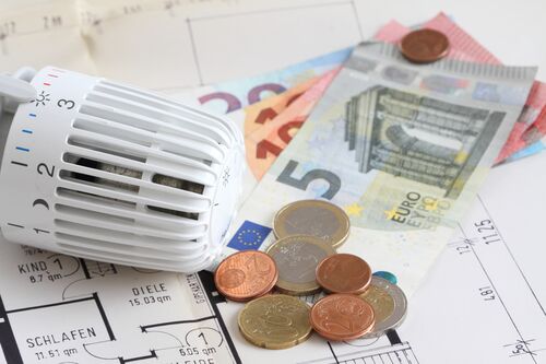 Euromünzen und Fünf-Euro-Schein liegen auf einem Wohnungsplan, daneben ein Heizthermostat