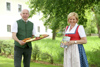 Landesrat Max Hiegelsberger steht mit einem Brett gefüllt mit heimischen Produkten neben Elfriede Schachinger, die ihr neues Buch in Händen hält, in einer Wiese.