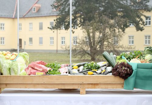 Auf einem Tisch eine Holzkiste mit Lebensmitteln: Äpfel, Kraut, Fleisch, Fische, Gurken, Salat. Im Hintergrund Baum und Kasernengebäude