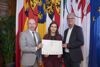 Landtagspräsident Max Hiegelsberger Dr.in Anna Obereder und Landtagsdirektor Dr. Wolfgang Steiner stehen nebeneinander und halten gemeinsam eine Urkunde, im Hintergrund Fahnen