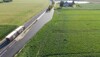  Blick aus der Vogelperspektive auf eine Straße mit einer Nebenfahrbahn bzw. Parkplatz inmitten grüner Felder. Auf der Straße fährt ein LKW,  im Hintergrund Bauernhof.