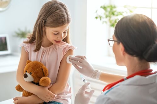 Ärztin tupft einem Mädchen, das einen Teddybär in den Armen hält, eine Impfstelle ab