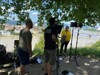 Monique Muhayimana steht im Schatten eines Baumes vor einer Kamera und zwei Scheinwerfern, ein Kameramann filmt, ein Techniker beobachtet, im Hintergrund die Donau, Linz-Urfahr und die Nibelungenbrücke