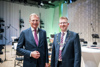 Landeshauptmann Mag. Thomas Stelzer und Rektor Martin Rummel stehen nebeneinander vor einer Bühne mit Einrichtung für ein Orchester, Stühle, Notenpulte, Mikrofone, Scheinwerfer