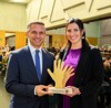 Landesrat Dr. Wolfgang Hattmannsdorfer und Mirna Jukic-Berger stehen nebeneinander in einem großen Saal mit Publikum und halten gemeinsam einen Preis aus Holz in Form zweier winkender Hände 