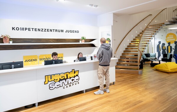 Blick auf den Innenraum und Front Desk des JugendService Campus in der Spittelwiese mit Mitarbeitenden des JugendService und einem Kunden.