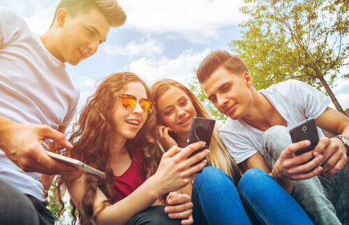 Vier Jugendliche sitzen nebeneinander mit Smartphones und schauen lächelnd gegenseitig auf die Displays.