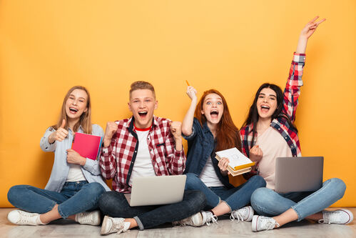 Vier junge Menschen sitzen nebeneinander und halten Laptops, Tablets und Notizblöcke in den Händen, mit einer Hand zeigen sie auf bzw. zeigen Daumen hoch