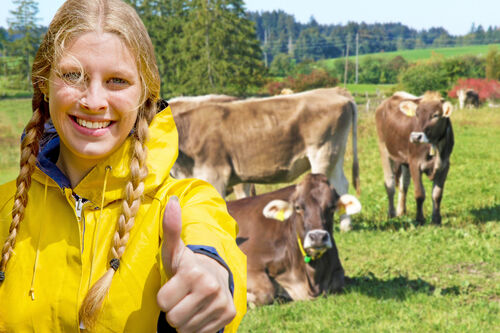 Junge Bäuerin deutet ok, im Hintergrund Kühe auf einer Weide