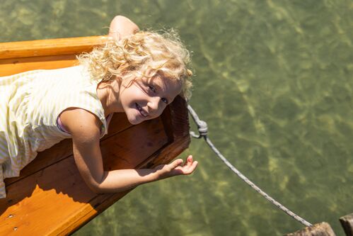 Ein lächelndes Mädchen liegt am Bug eines Ruderbootes aus Holz auf einem See und zeigt mit der Hand ins klare Wasser