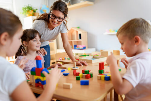 Kinder an einem Tisch spielen mit Bauklötzen, Pädagogin stehend neben den Kindern