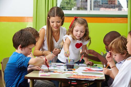 Kindergartenpädagogin sitzt mit sechs Kindern an einem niedrigen Tisch, die Kinder malen mit Wasserfarben