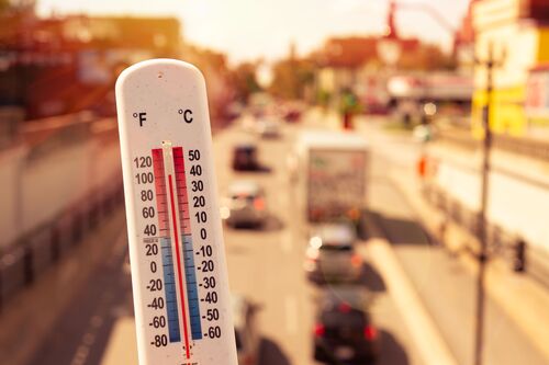 Thermometer zeigt fast 40 Grad Celsius an, im Hintergrund städtisches Gebiet mit Häusern, Straßen, Autoverkehr