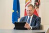 Landeshauptmann Mag. Thomas Stelzer sitzt an einem Schreibtisch und tippt auf ein Tablet, im Hintergrund Europa-, Oberösterreich- und Österreich-Fahne