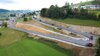 Straßenbaumaschinen auf einem frisch asphaltierten Serpentinen-Straßenstück, Ansicht auf eine Ortschaft in einer hügeligen Landschaft