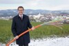 Landesrat Markus Achleitner hält ein großes Stück Glasfaserkabel in Händen, im Hintergrund eine kleine Stadt, Berge, Feld