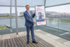 LR Markus Achleitner steht vor einer Glasfront und Hält ein vergrößertes Smartphoneplakat in der Hand, dahinter die Donau und die Stadt Linz.
