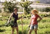 Michaela Jancsy und Reingard Prohaska stehen nebeneinander in einem Feld mit Kräutern und Gemüse, im Hintergrund ein großes Glashaus und Bäume