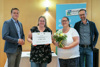 Landesrat Stefan Kaineder, Anita Wiesinger mit Gutschein, Gudrun Bumberger mit Blumenstrauß und Norbert Rainer stehen nebeneinander in einem Raum, hinter ihnen Rollplakat mit Beschriftung Klimabündnis Oberösterreich