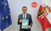 Wirtschafts- und Europa-Landesrat Markus Achleitner mit dem Interreg-Programm in den Händen, stehend zwischen der Oberösterreich-Fahne und der EU-Fahne.