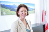 Landesrätin Michaela Langer-Weninger steht in einem Büro, im Hintergrund großes Foto an der Wand mit einem Landschaftsmotiv und Oberösterreich-Fahne
